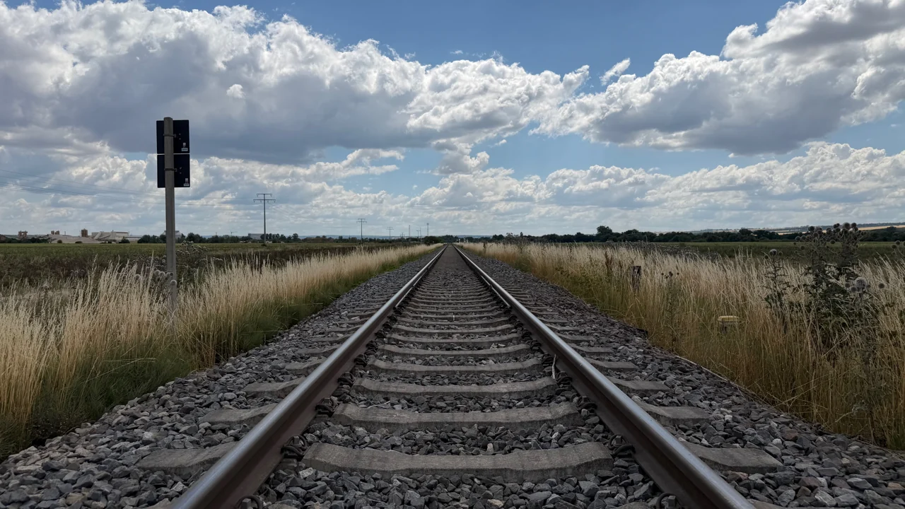 Ein gerades Bahngleis führt zwischen Feldern zum Horizont; daneben stehen Masten und ein Signal.